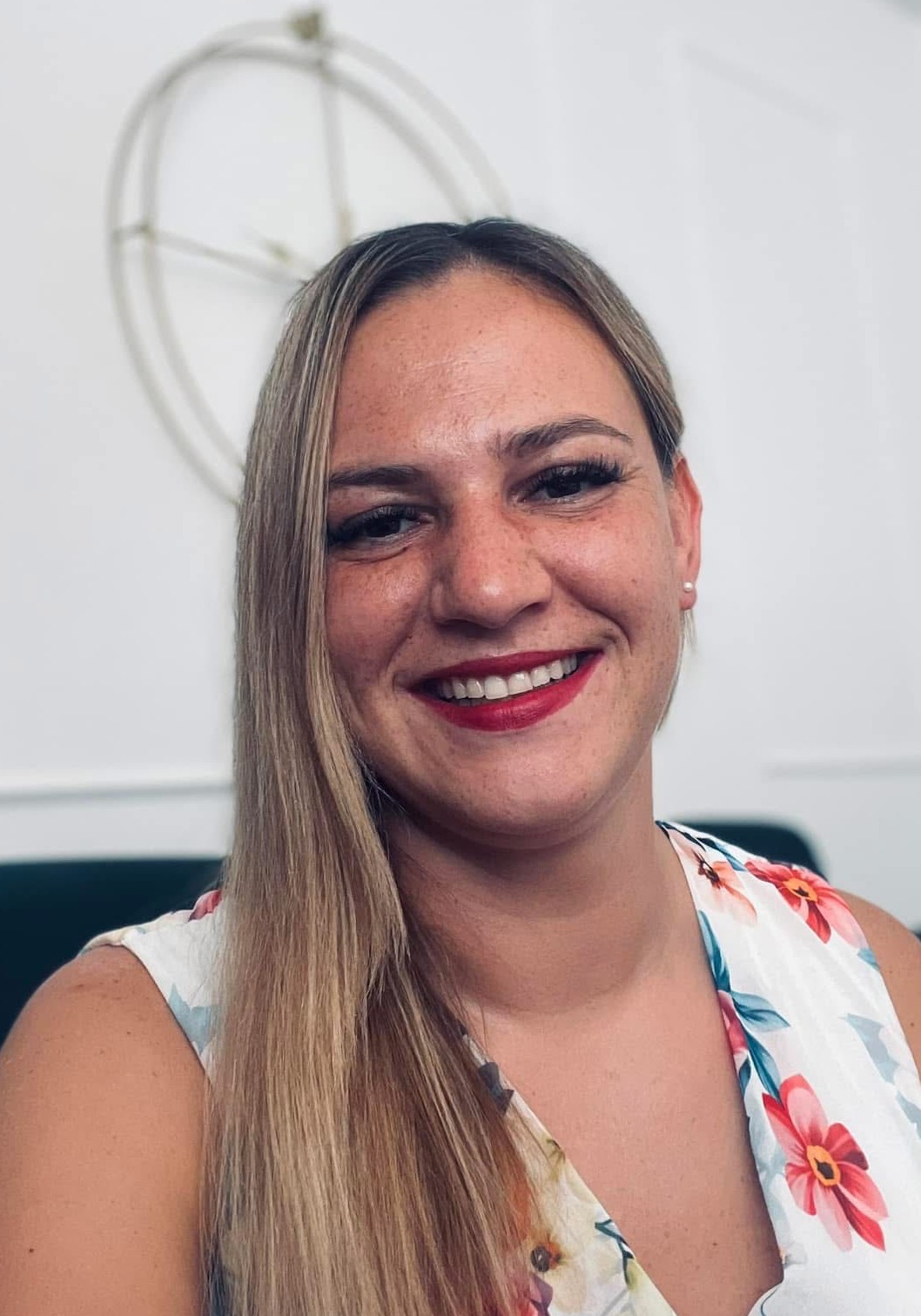 Uśmiechnięta kobieta z blond włosami i czerwonymi ustami, ubrana w bluzkę w kwiaty, na tle białej ściany z dekoracją.