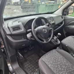 Wnętrze samochodu Opel Combo z widocznymi detalami deski rozdzielczej, kierownicy z logo Opel i wzorzystymi siedzeniami. Na przedniej szybie widoczne krople deszczu.