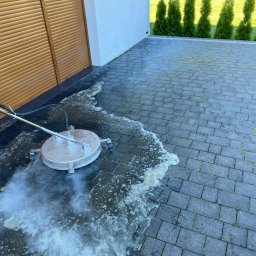 Czyścioch Usługi Sprzątające - Czyszczenie kostki brukowej myjką ciśnieniową z rotacyjną dyszą. Widoczna piana i para wodna, częściowo umyta powierzchnia. Tło: budynek z brązową bramą i zielone tuje.