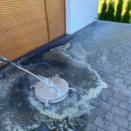 Czyścioch Usługi Sprzątające - Mycie kostki brukowej przy użyciu specjalistycznej maszyny. Widoczne efekty czyszczenia, woda i piana. W tle brama garażowa i tuje.
