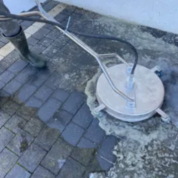 Czyszczenie kostki brukowej myjką ciśnieniową z okrągłą dyszą. Widoczne różnice w czystości kostki. Osoba w zielonych kaloszach obsługuje urządzenie.