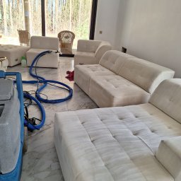 Czyścioch Usługi Sprzątające - Pranie narożnika modułowego w salonie z widokiem na las. Widoczny sprzęt do czyszczenia tapicerki i efekt prania na jasnym materiale. Marmurowa podłoga.
