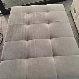 Czyścioch Usługi Sprzątające - Kwadratowy, jasny puf z widocznymi śladami po praniu tapicerki, umieszczony na marmurowej podłodze przy oknie. Część pufa jest poza kadrem.