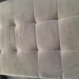 Widok z g&oacute;ry na beżowy, tapicerowany puf z widocznymi plamami i zabrudzeniami, ukazujący potrzebę profesjonalnego czyszczenia tapicerki meblowej.