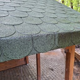 Czyścioch Usługi Sprzątające - Zielony dach z gontu bitumicznego, lekko wilgotny po deszczu, z widocznymi kroplami wody. Konstrukcja drewniana z kostką brukową w tle i zielenią drzew.