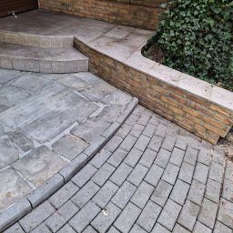 Czyścioch Usługi Sprzątające - Szara kostka brukowa ułożona przy schodach i murku z cegły, obok zielony bluszcz. Widoczne drobne zabrudzenia na kostce.