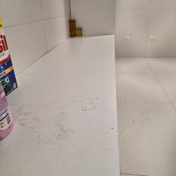 Czyścioch Usługi Sprzątające - Brudny, biały blat kuchenny z plamami i resztkami, obok butelka płynu i proszek do prania. W tle białe płytki i podłoga w podobnym stylu.