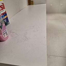 Czyścioch Usługi Sprzątające - Brudny blat w łazience z resztkami proszku i płynu do prania. Widoczne płytki ścienne i podłogowe o prążkowanej fakturze oraz pojemniki na blacie.