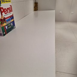 Czyścioch Usługi Sprzątające - Półka z detergentem Persil w nowoczesnej łazience z białymi płytkami i widocznymi gniazdkami elektrycznymi. Minimalistyczny design i czystość.