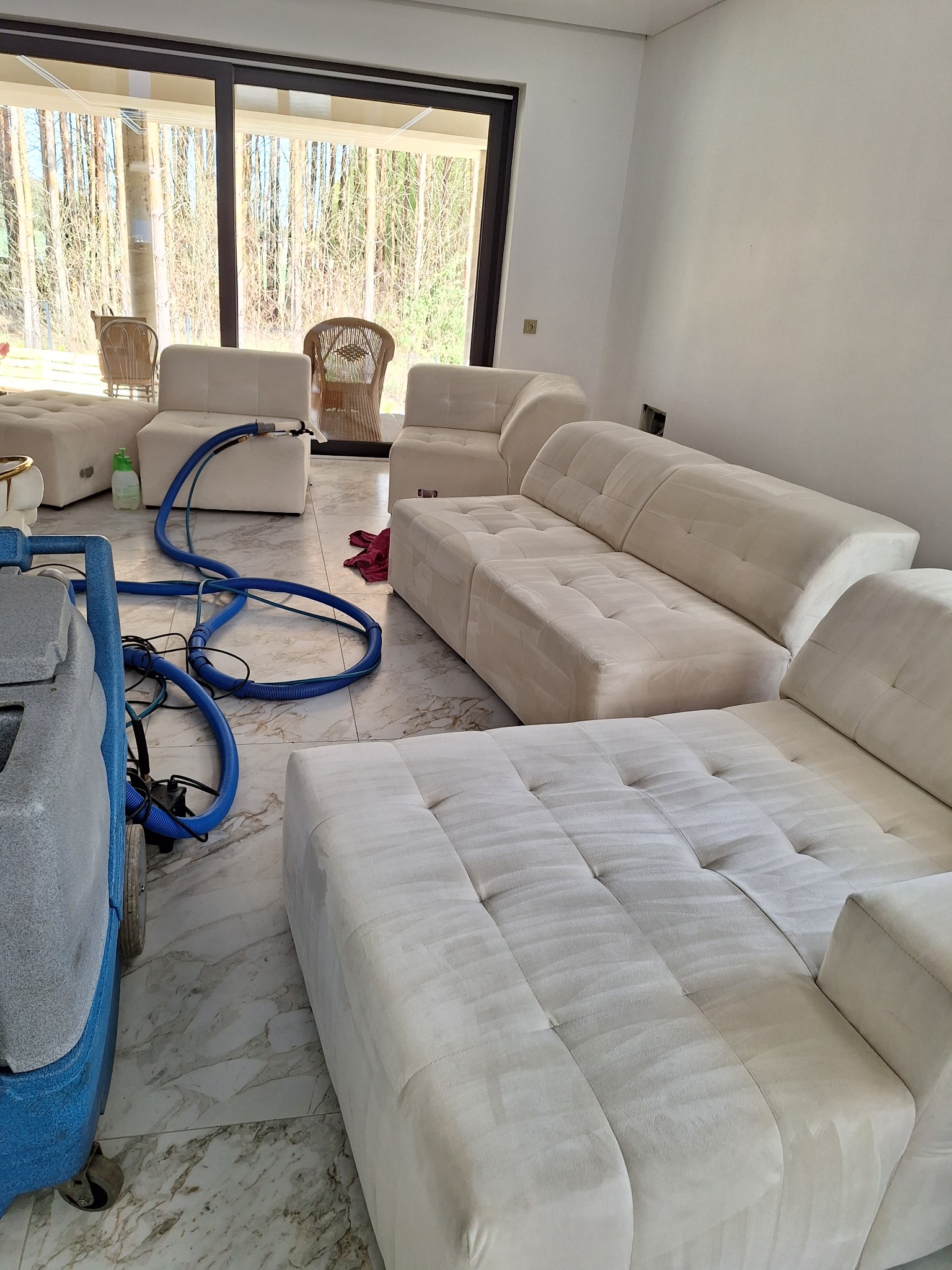 Pranie narożnika modułowego w salonie z widokiem na las. Widoczny sprzęt do czyszczenia tapicerki i efekt prania na jasnym materiale. Marmurowa podłoga.