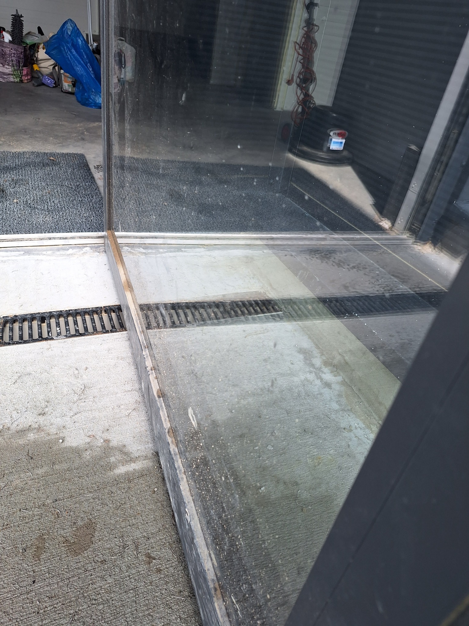 Brudne okno po czyszczeniu z widocznymi smugami i zaciekami na szklanej powierzchni, zarys kratki ściekowej na betonowej posadzce.