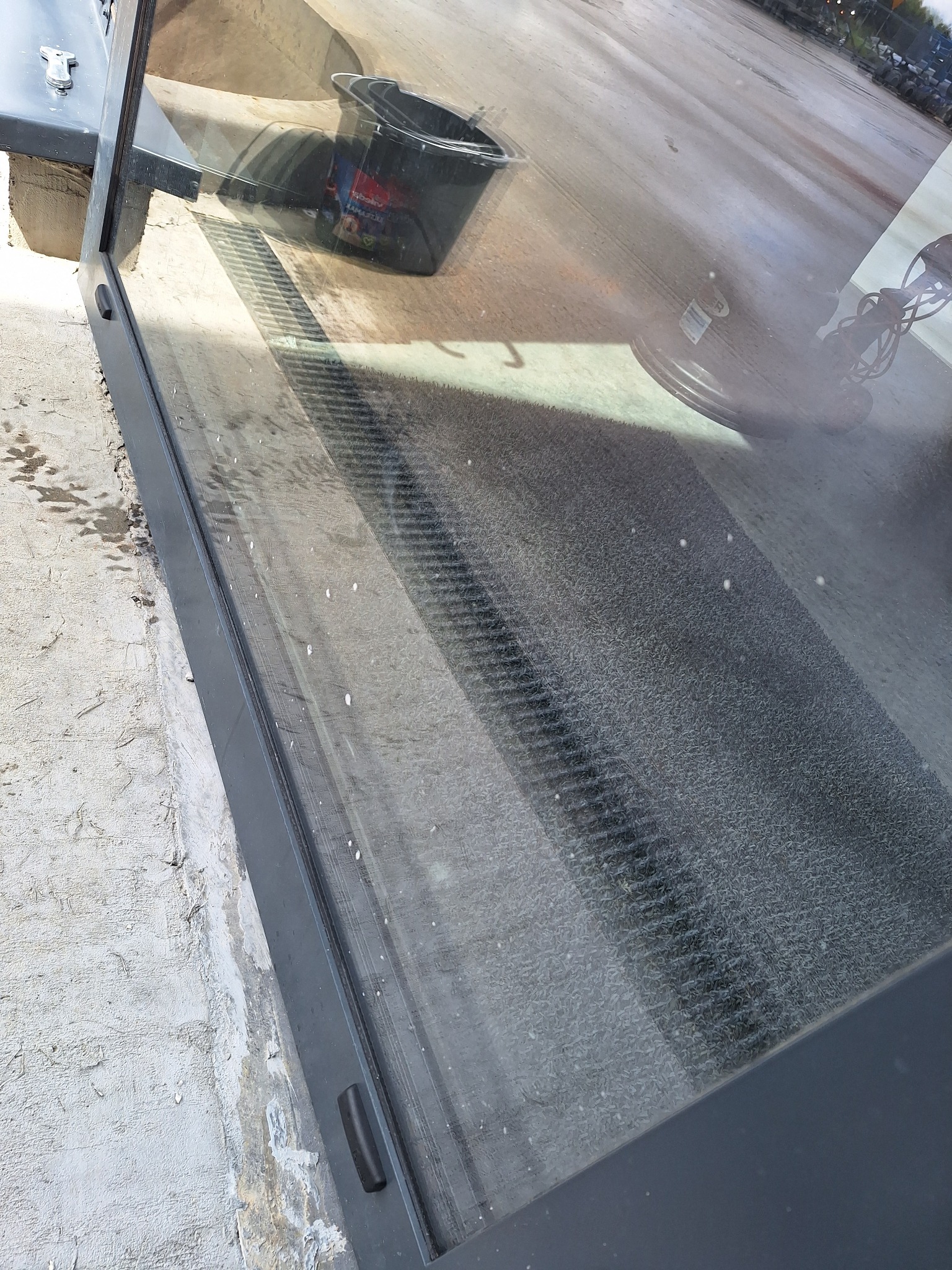 Brudna szyba okienna z widocznymi smugami i plamami. W tle wiadro i sprzęt do mycia. Widoczne ślady po umyciu fragmentu szyby, co sugeruje trwający proces czyszczenia.