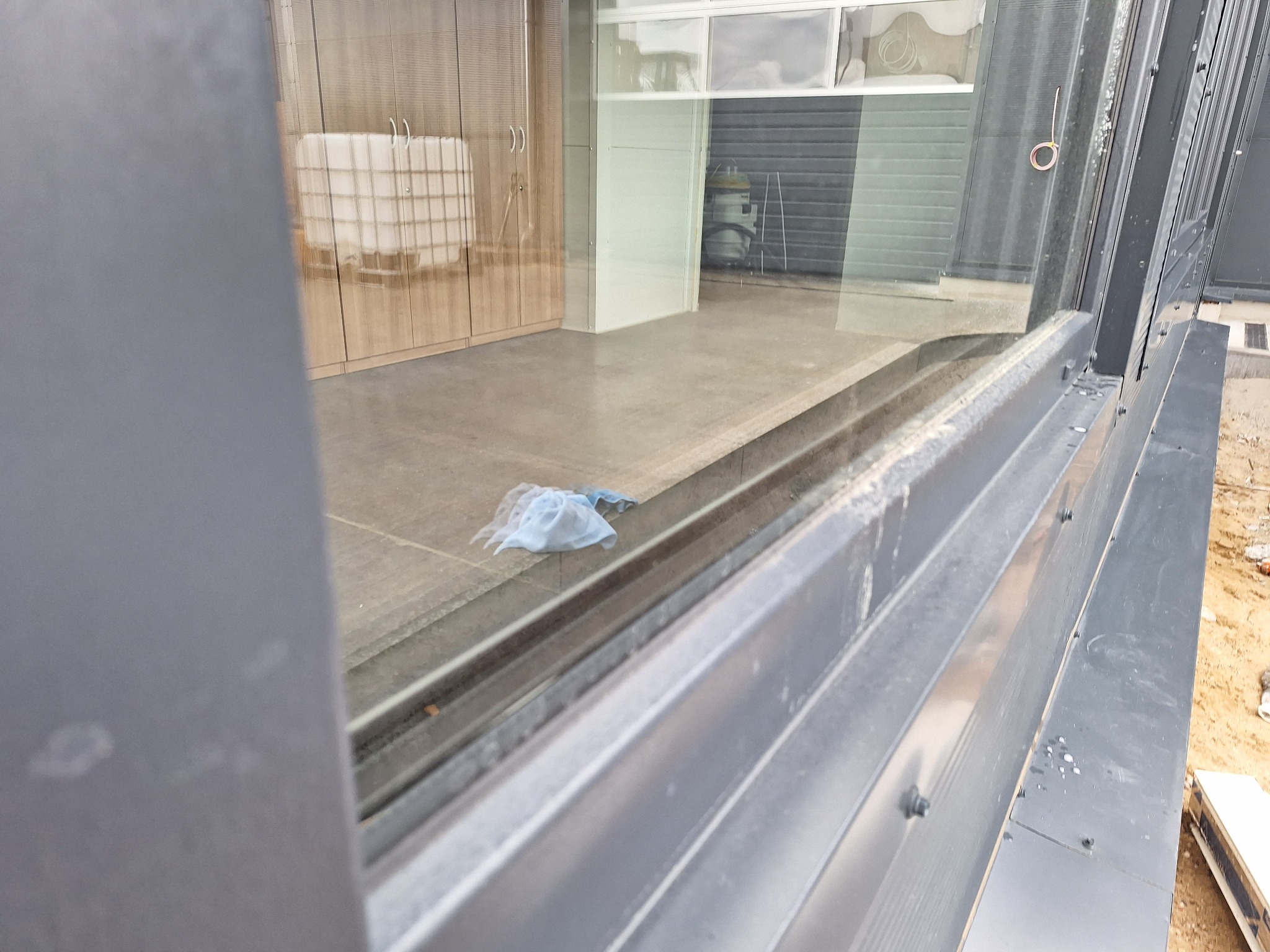 Widok przez brudne okno na posadzkę z niebieską szmatką i meble w tle. Okno z szarymi ramami, widoczne ślady po myciu. Perspektywa z dołu.