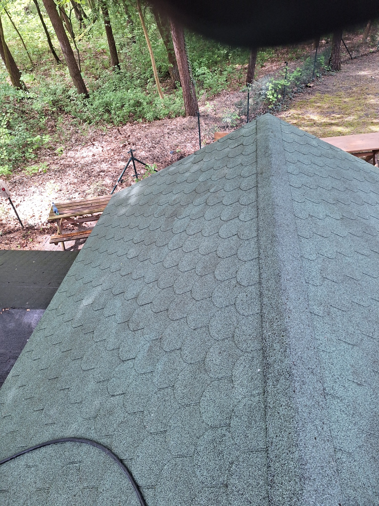 Z bliska: zielony dach altany pokryty bitumiczną dachówką na tle leśnego otoczenia. Widoczny fragment stołu piknikowego i kabel na dachu.