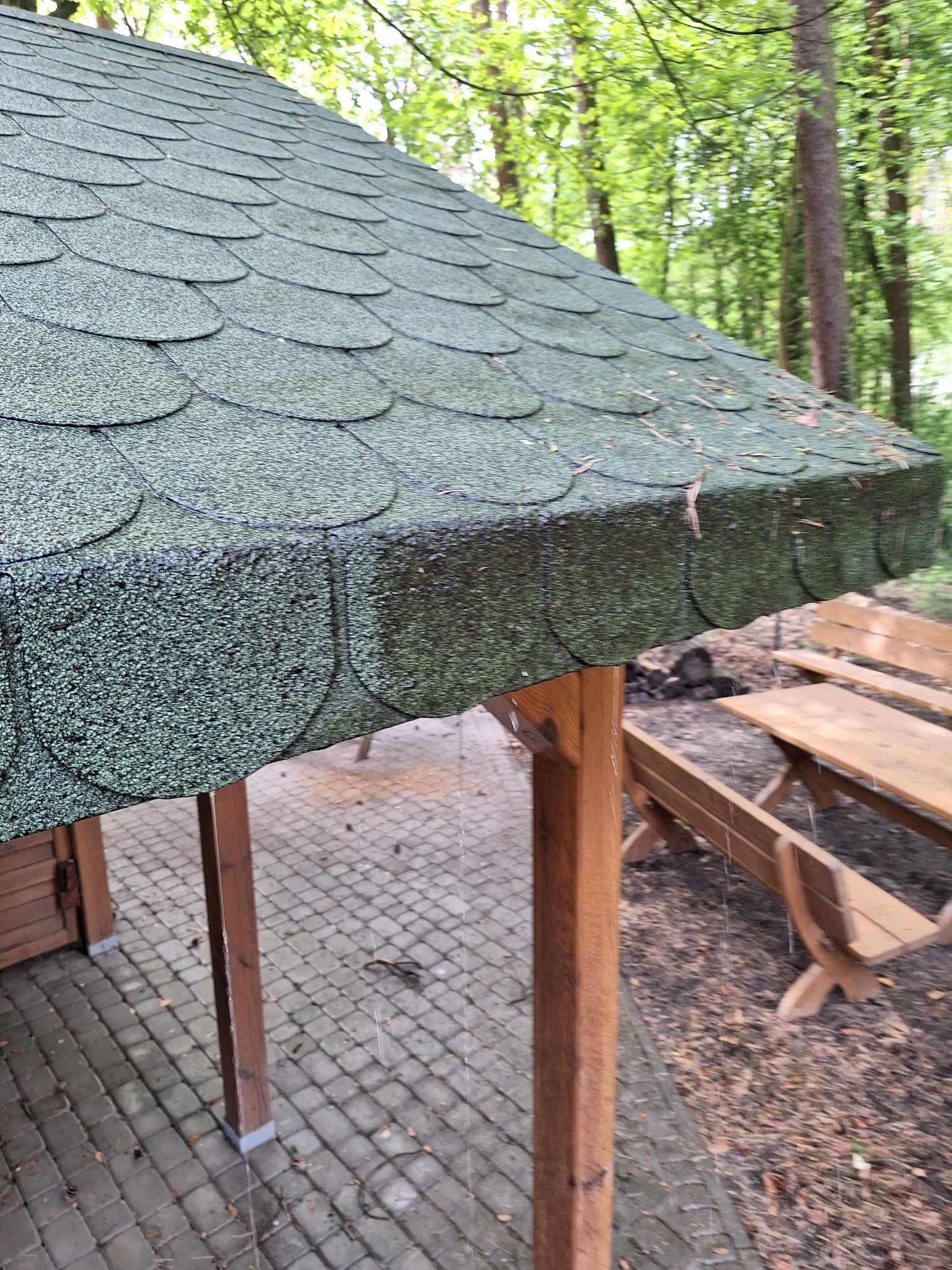 Zielony dach altany pokryty gontem bitumicznym, widok z dołu, fragment drewnianej konstrukcji i ławki piknikowej w tle, otoczenie leśne.
