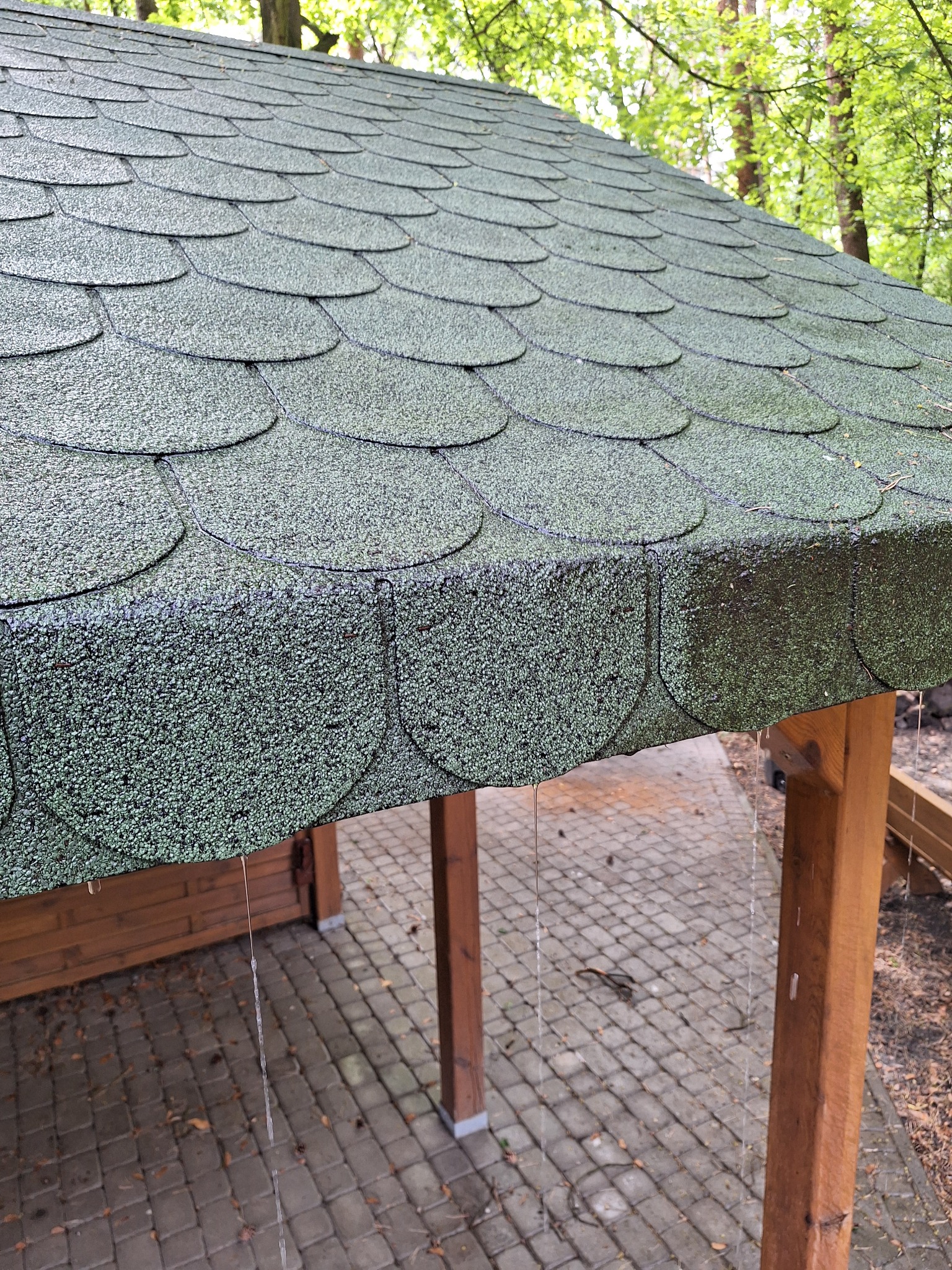 Zielony dach z gontu bitumicznego, lekko wilgotny po deszczu, z widocznymi kroplami wody. Konstrukcja drewniana z kostką brukową w tle i zielenią drzew.