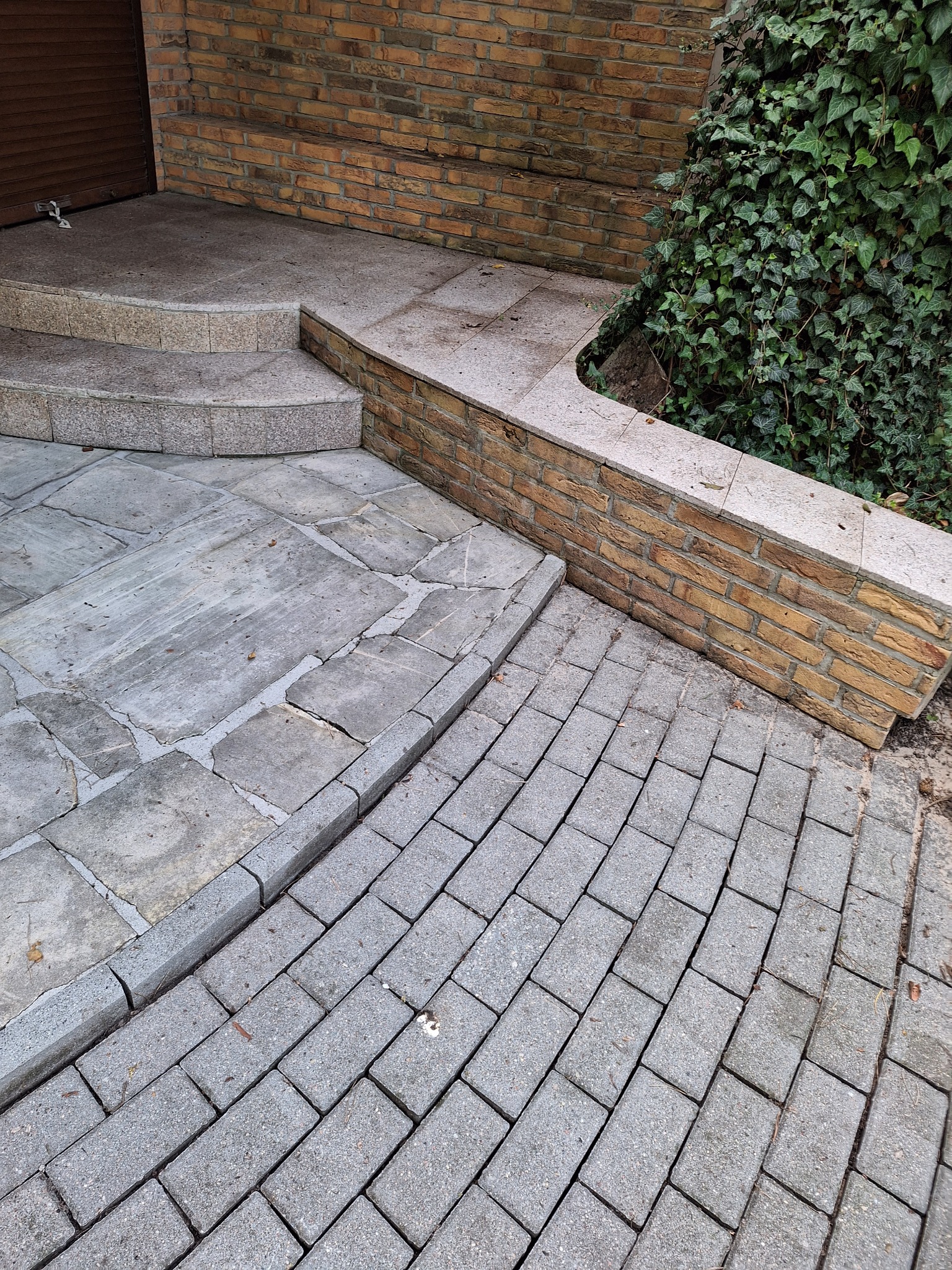 Szara kostka brukowa ułożona przy schodach i murku z cegły, obok zielony bluszcz. Widoczne drobne zabrudzenia na kostce.