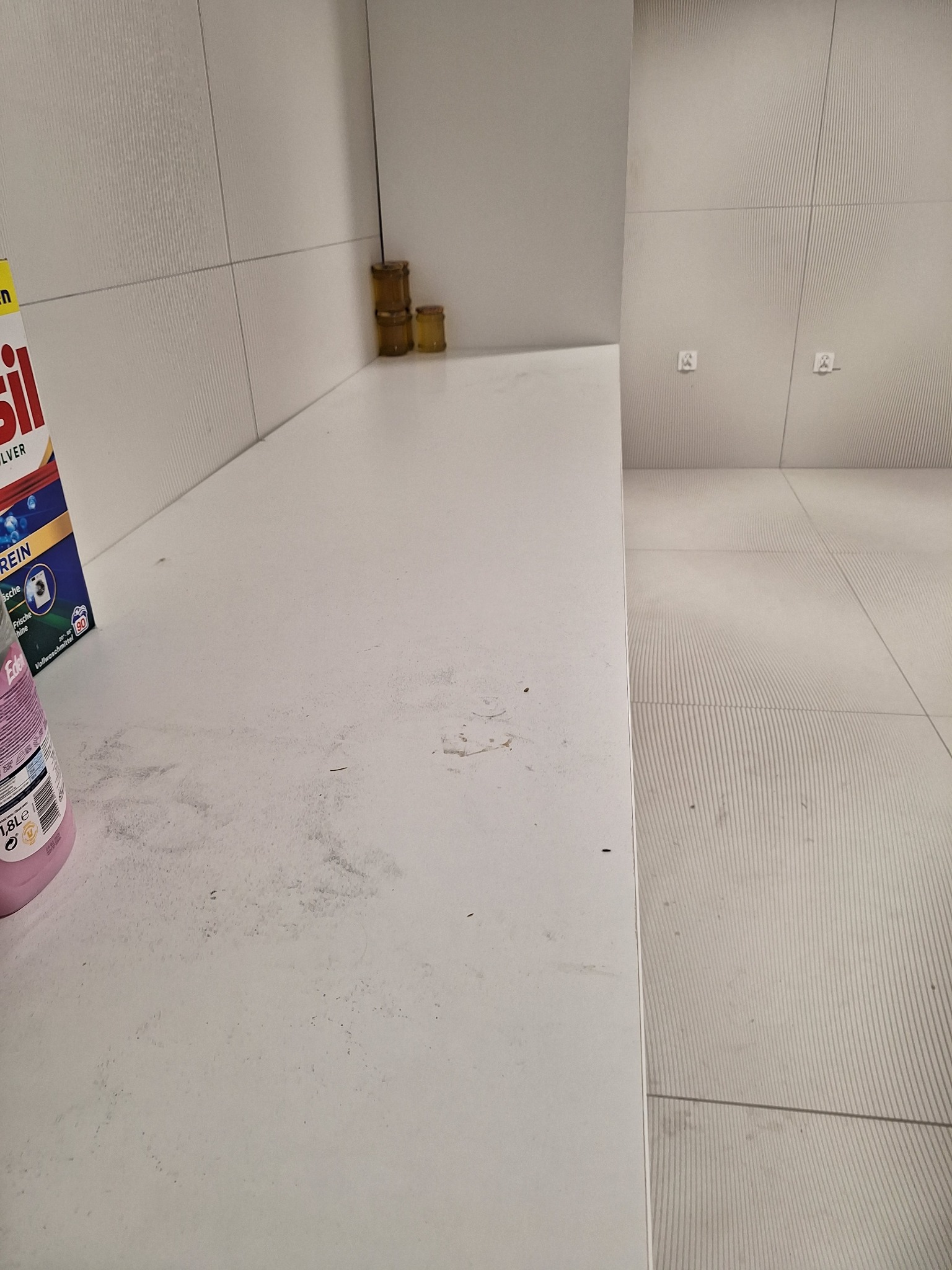 Brudny, biały blat kuchenny z plamami i resztkami, obok butelka płynu i proszek do prania. W tle białe płytki i podłoga w podobnym stylu.