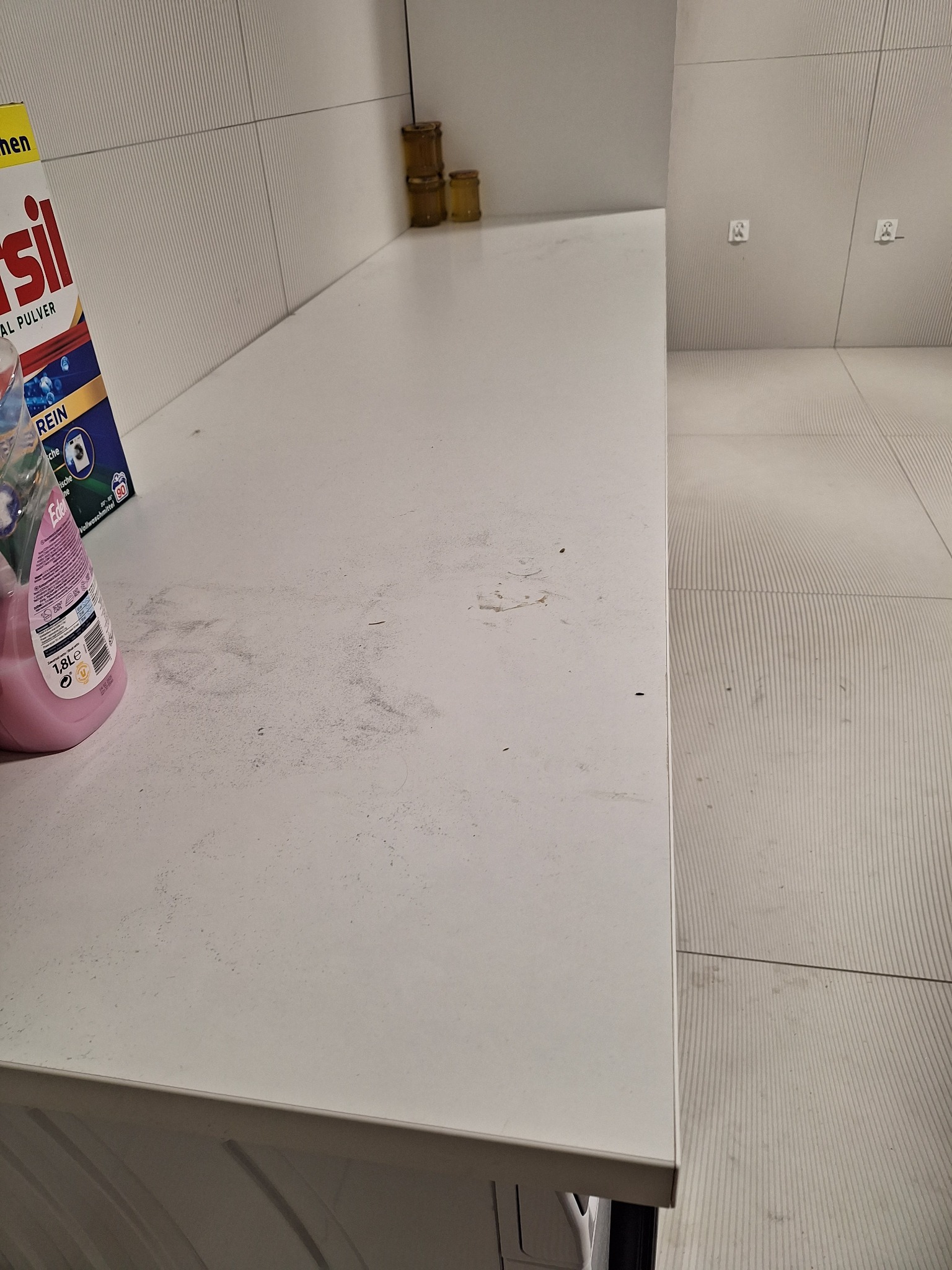 Brudny blat w łazience z resztkami proszku i płynu do prania. Widoczne płytki ścienne i podłogowe o prążkowanej fakturze oraz pojemniki na blacie.