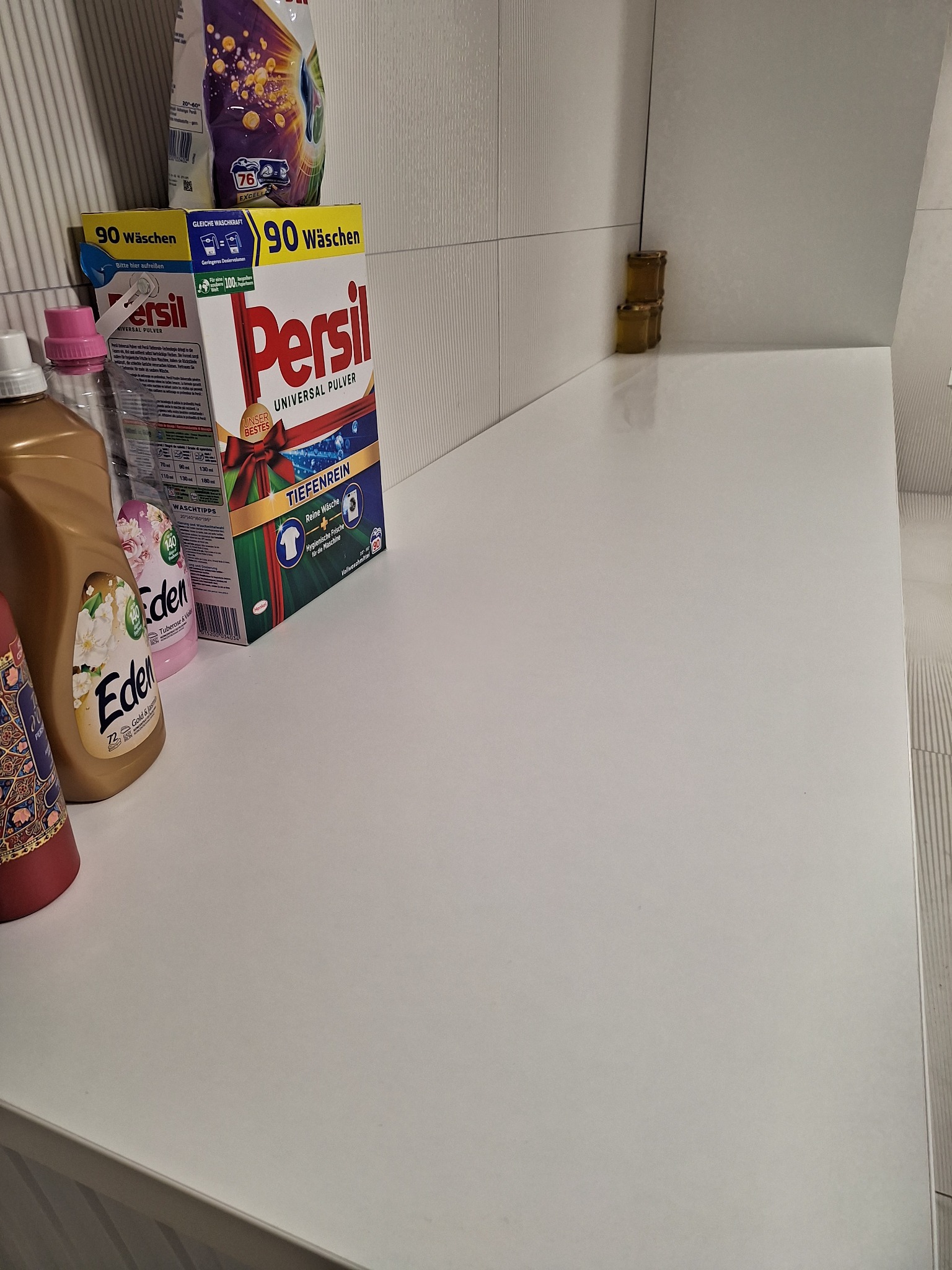 Środki czystości: płyny i proszki do prania Eden i Persil na białym blacie w łazience z białymi płytkami. Częściowe ujęcie produktów.