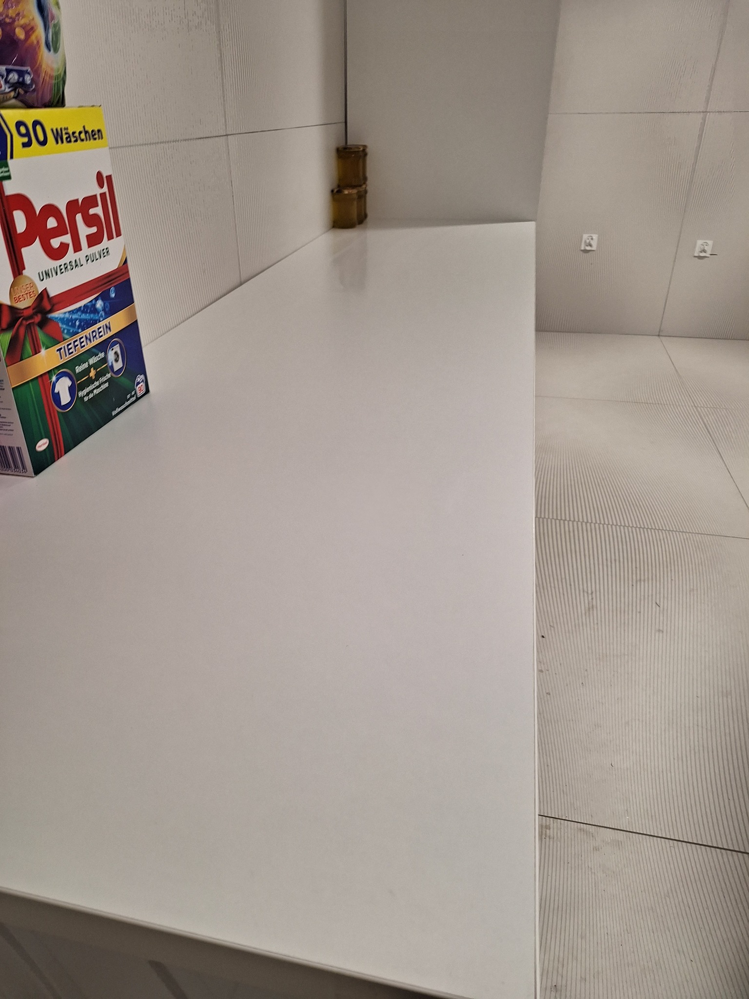 Półka z detergentem Persil w nowoczesnej łazience z białymi płytkami i widocznymi gniazdkami elektrycznymi. Minimalistyczny design i czystość.