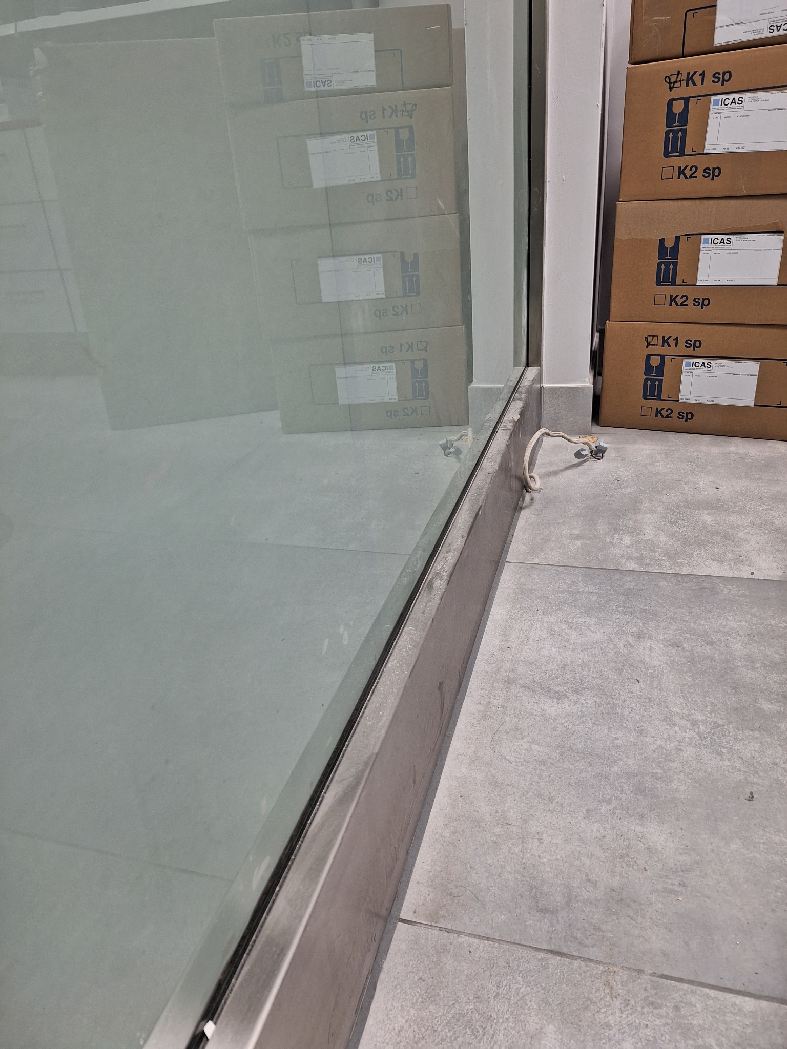 Umyta szyba z widocznymi kartonami w tle. Widoczne ślady po czyszczeniu na podłodze. Kabel elektryczny wystaje spod szyby. Szary kolor podłogi.