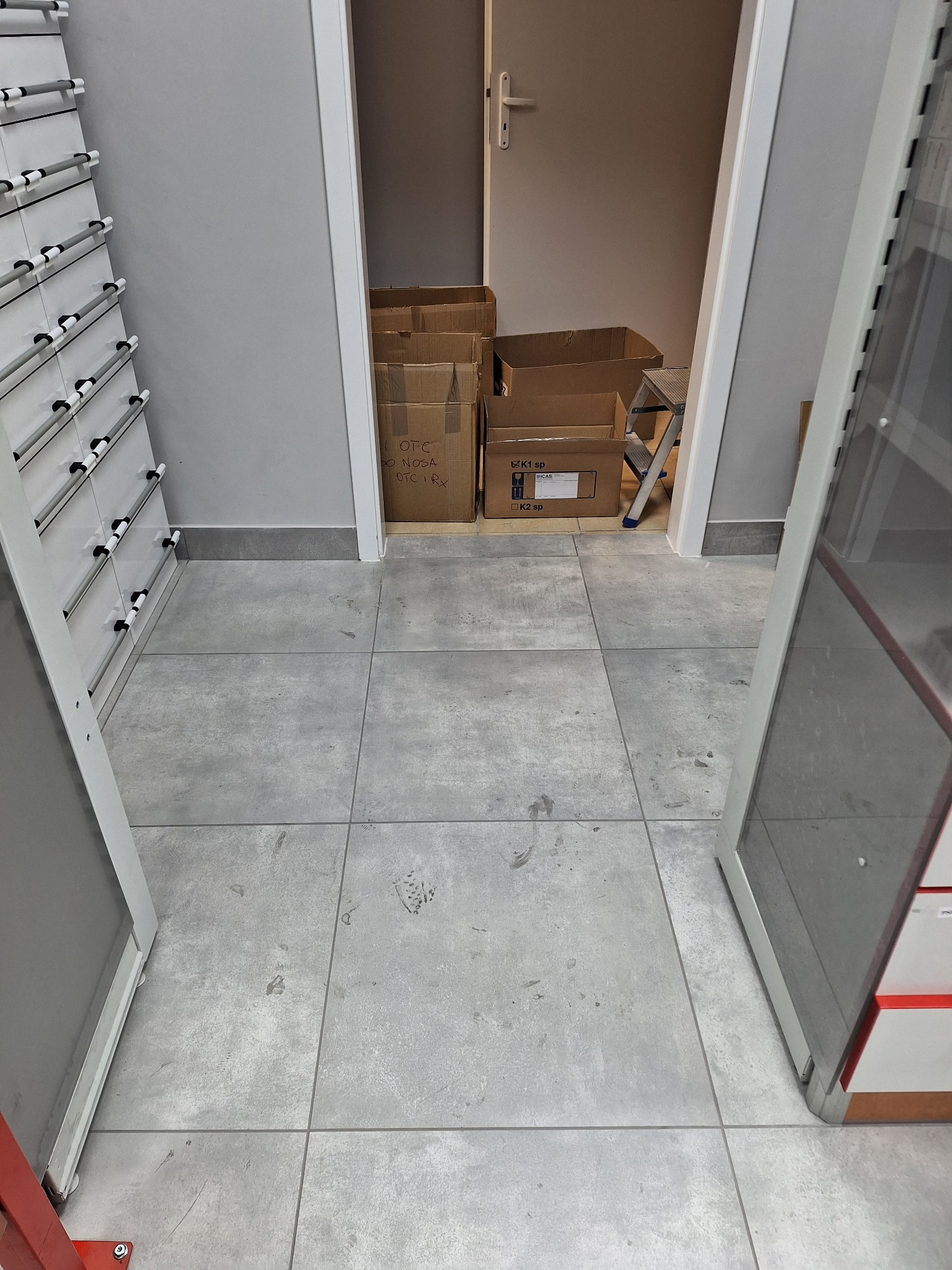 Szare płytki podłogowe z widocznymi śladami zabrudzeń, kartony w tle, fragment regału i drzwi. Ujęcie z góry, ukazujące potencjalny obszar wymagający sprzątania.