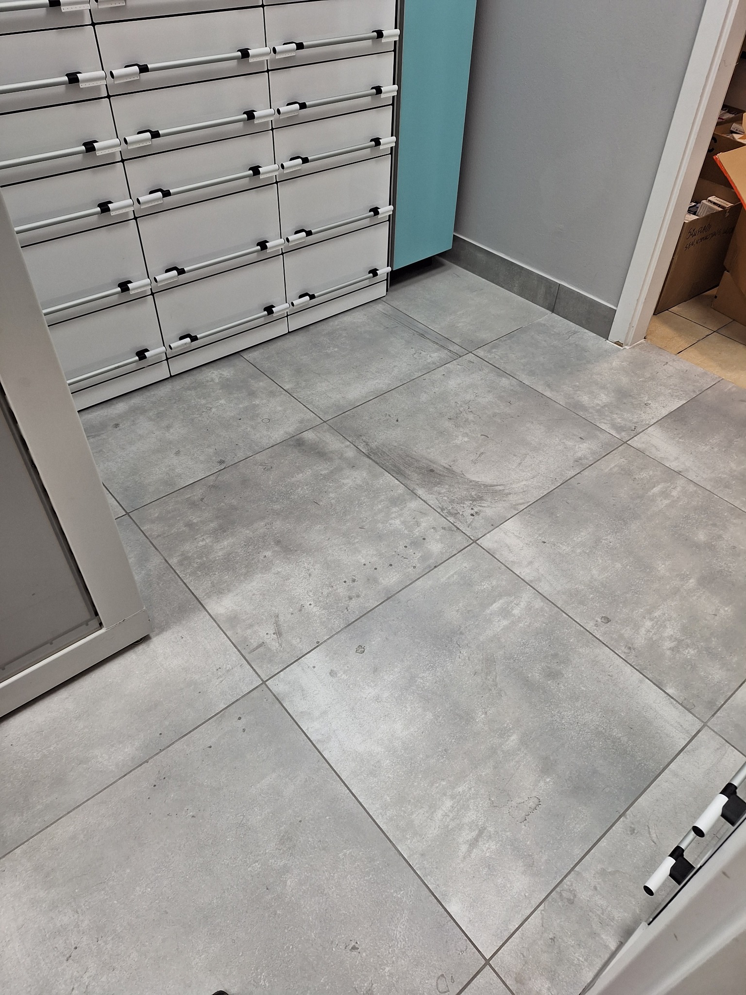 Szare płytki podłogowe z widocznymi śladami zabrudzeń, częściowo umyte. W tle białe szafki z szufladami i fragment ściany. Widoczne niedociągnięcia w czyszczeniu.
