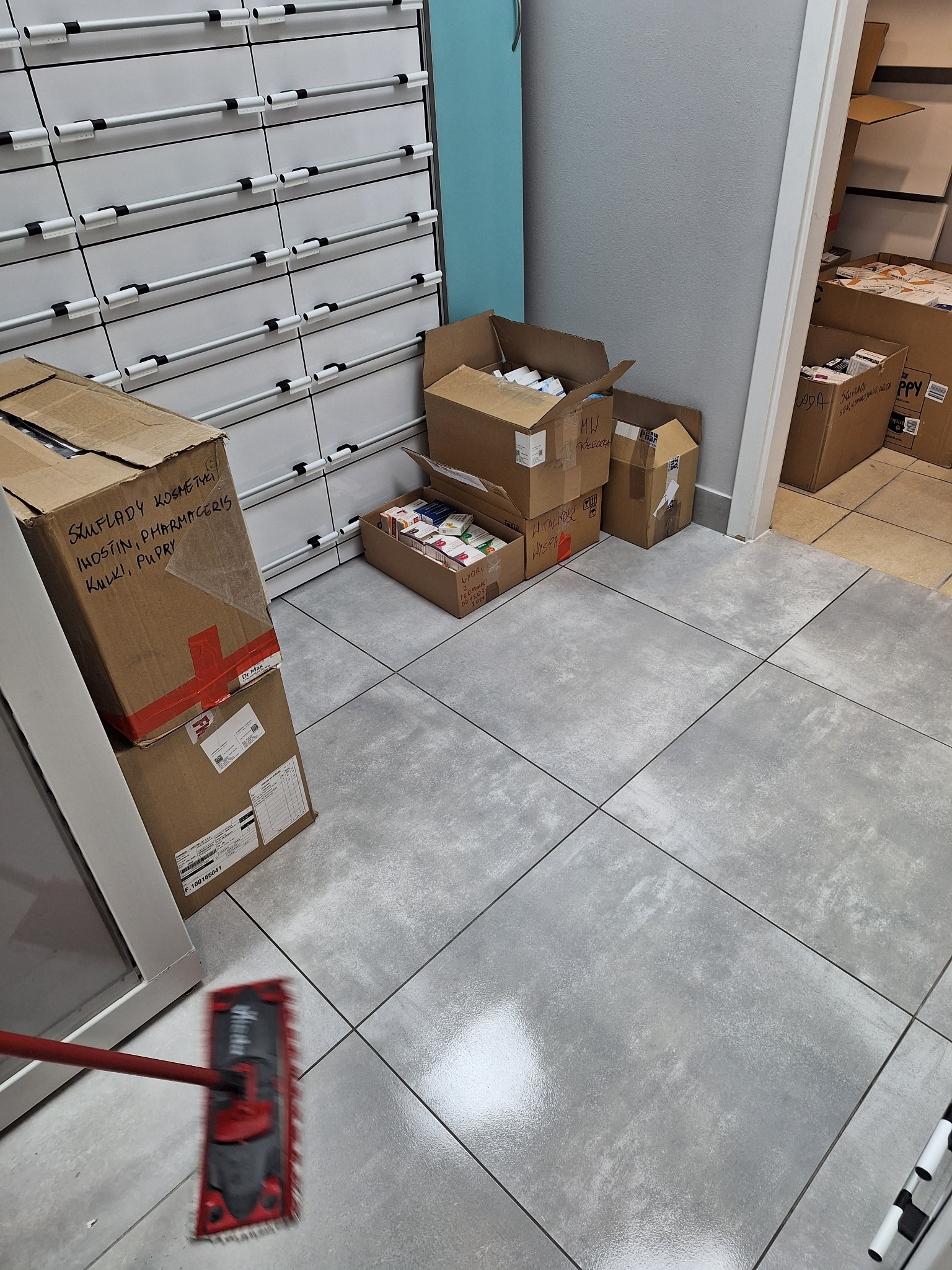 Podłoga w pomieszczeniu aptecznym w trakcie sprzątania, widoczne kartony z lekami i mop. Ujęcie z góry, z perspektywy osoby sprzątającej.