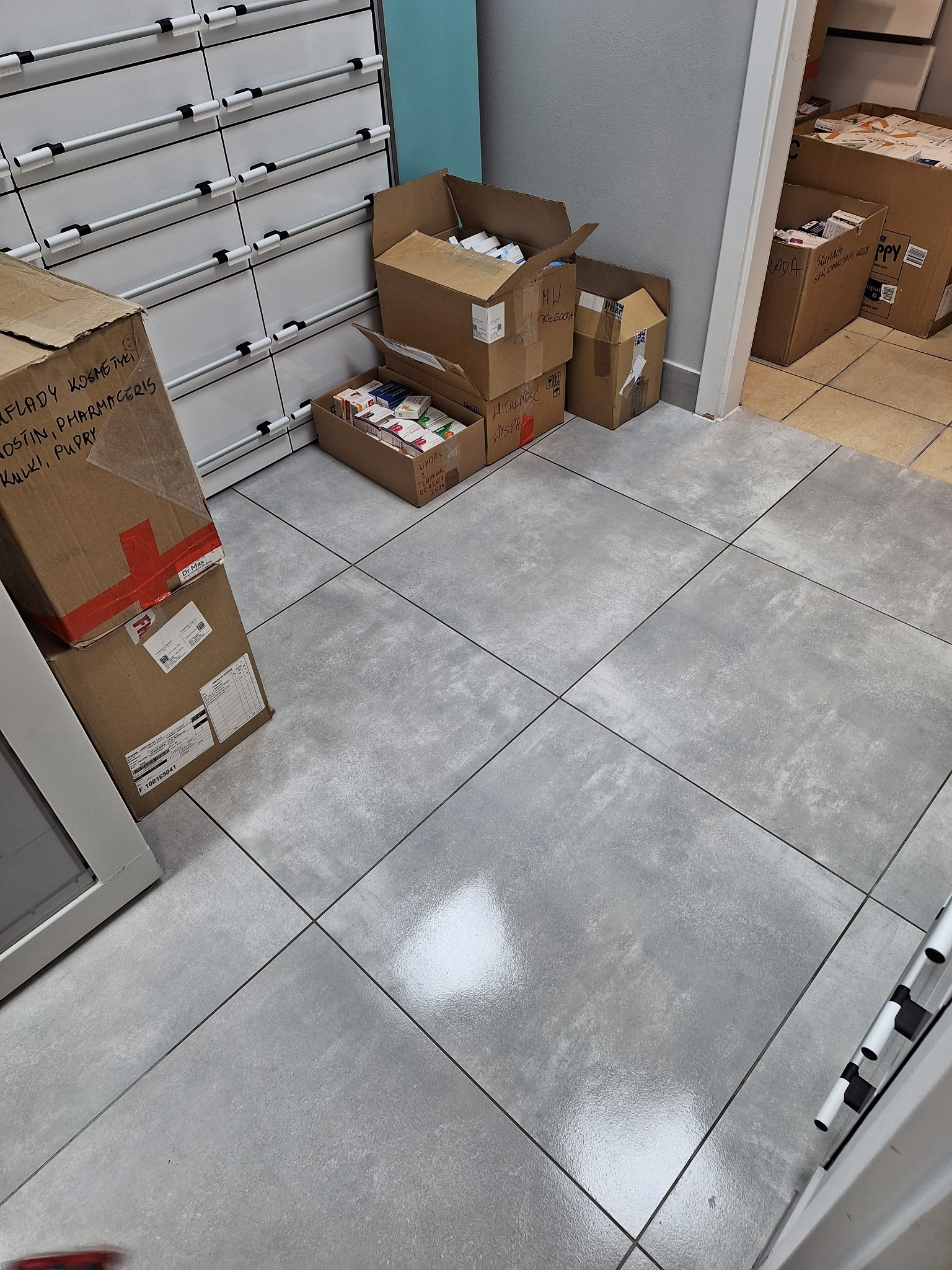 Kartonowe pudła z lekami i kosmetykami ustawione przy białych szafkach aptecznych i szarej ścianie. Część pudeł otwartych, widoczne opakowania leków.