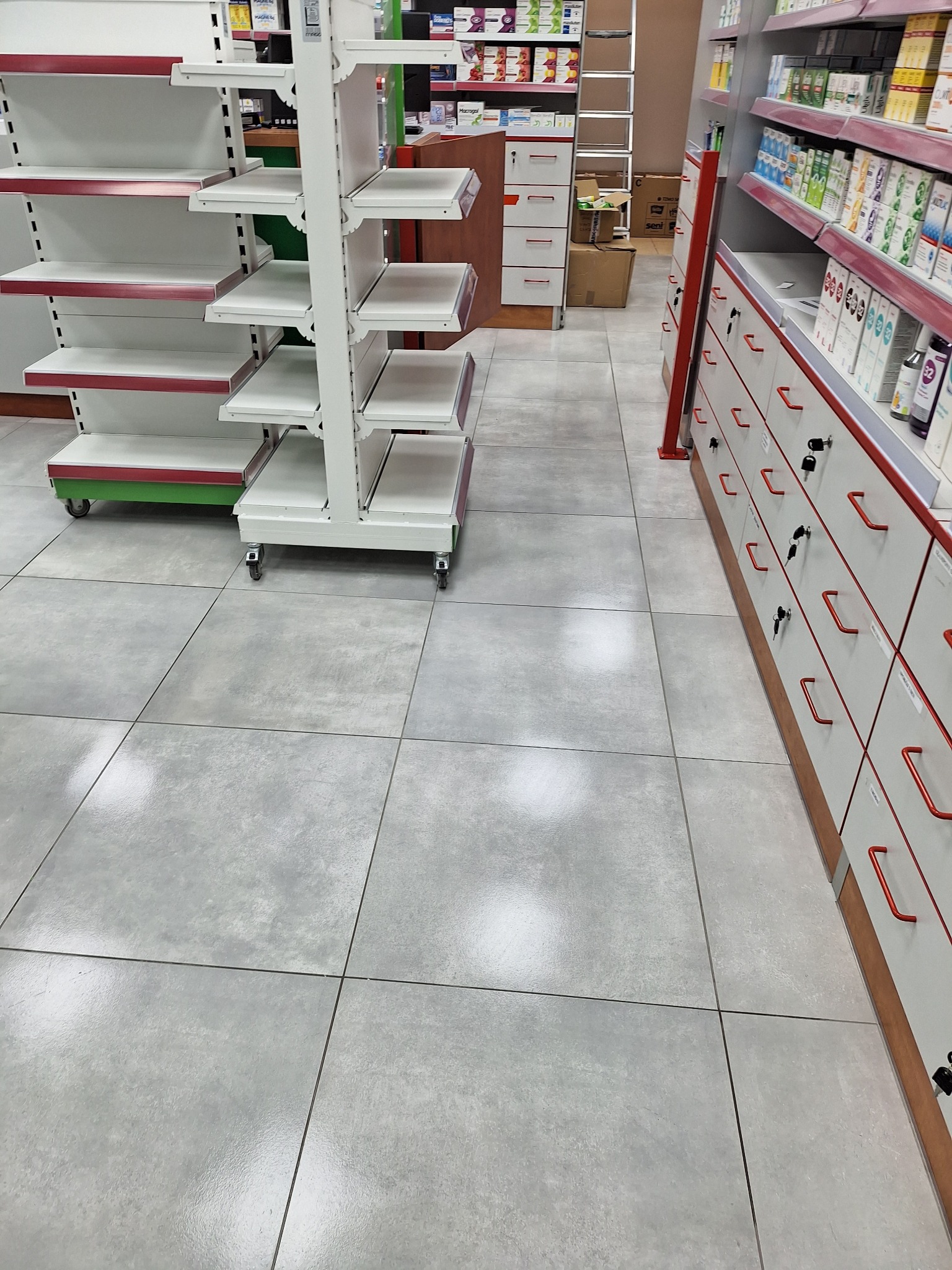 Wnętrze apteki z szarymi płytkami podłogowymi, białymi regałami na kółkach i szafkami z szufladami oraz lekami. Widok na korytarz apteczny.