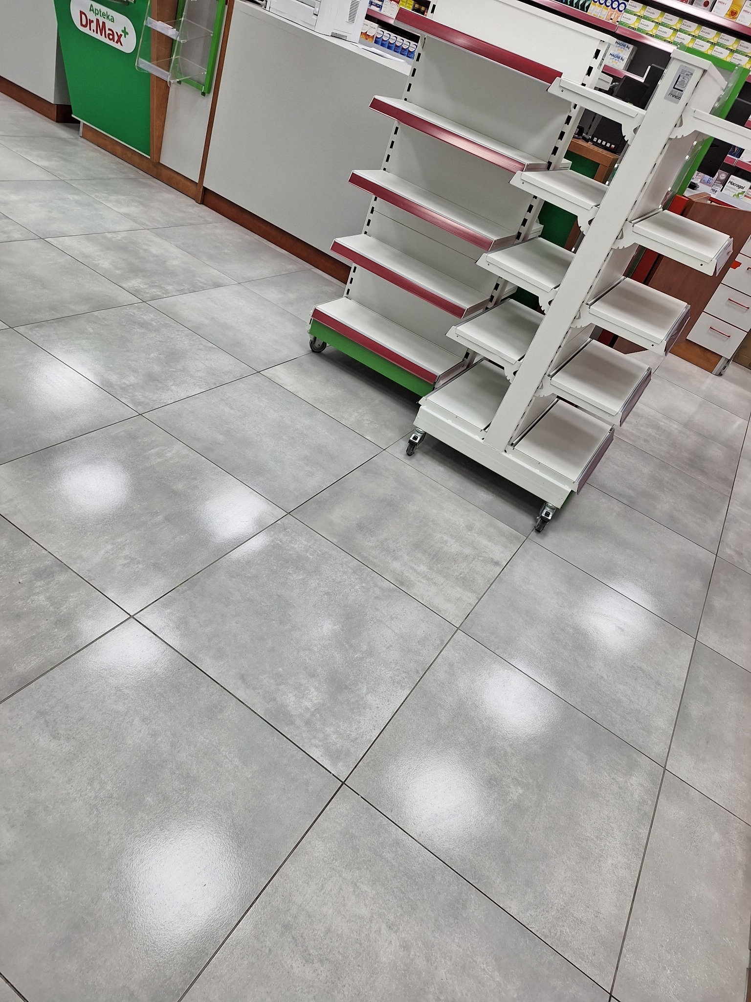 Półki sklepowe na kółkach na tle szarych płytek podłogowych w aptece Dr.Max. Widok z góry, lekko ukośny. Puste regały, gotowe do ekspozycji towaru.