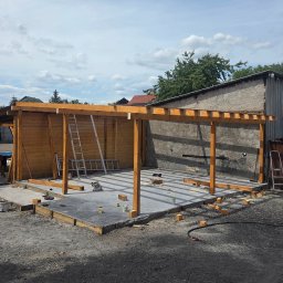 Riko Kompleksowe remonty - Drewniana konstrukcja zadaszenia w trakcie budowy, widoczne słupy, belki, podłoże betonowe, drabina oparta o ścianę. Elementy konstrukcyjne w kolorze jasnego drewna.