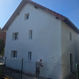 Riko Kompleksowe remonty - Biały dom jednorodzinny z czerwoną dachówką w trakcie malowania elewacji przez pracownika, widok zza siatki ogrodzeniowej w słoneczny dzień.