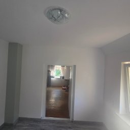 Riko Kompleksowe remonty - Wnętrze po remoncie: białe ściany, szara kolumna, widok na sąsiedni pokój z drewnianą podłogą, nowoczesna lampa sufitowa, okno z widokiem na zewnątrz.