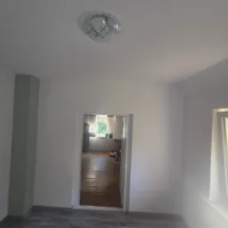 Wnętrze po remoncie: białe ściany, szara kolumna, widok na sąsiedni pokój z drewnianą podłogą, nowoczesna lampa sufitowa, okno z widokiem na zewnątrz.