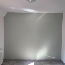 Riko Kompleksowe remonty - Pomieszczenie po malowaniu: szara ściana z delikatnym połyskiem światła, białe ściany boczne, grzejnik i panele podłogowe. Widok na odnowione wnętrze.