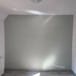 Pomieszczenie po malowaniu: szara ściana z delikatnym połyskiem światła, białe ściany boczne, grzejnik i panele podłogowe. Widok na odnowione wnętrze.