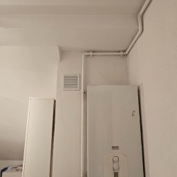 Riko Kompleksowe remonty - Biały piec gazowy zamontowany na ścianie w rogu pomieszczenia, z widocznymi rurami i kratką wentylacyjną. Obok szafka, a w tle skos dachu.