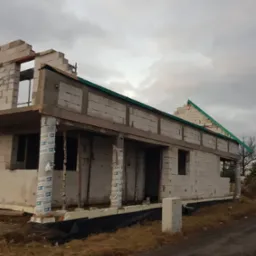 Surowy stan domu w budowie z bloczków, z widocznymi elementami konstrukcyjnymi dachu i izolacją kolumn. Widok na szary budynek w trakcie wznoszenia.