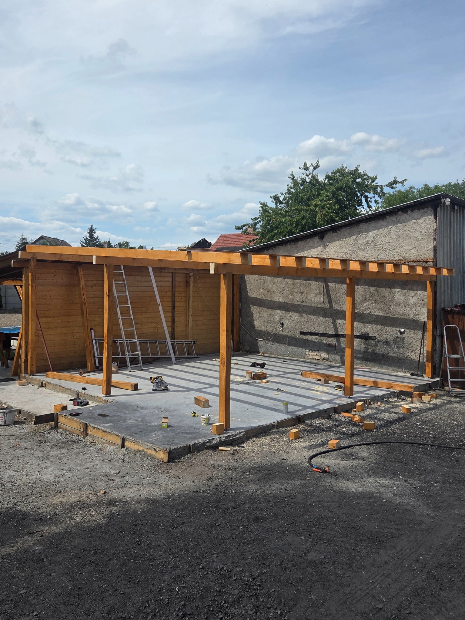 Drewniana konstrukcja zadaszenia w trakcie budowy, widoczne słupy, belki, podłoże betonowe, drabina oparta o ścianę. Elementy konstrukcyjne w kolorze jasnego drewna.