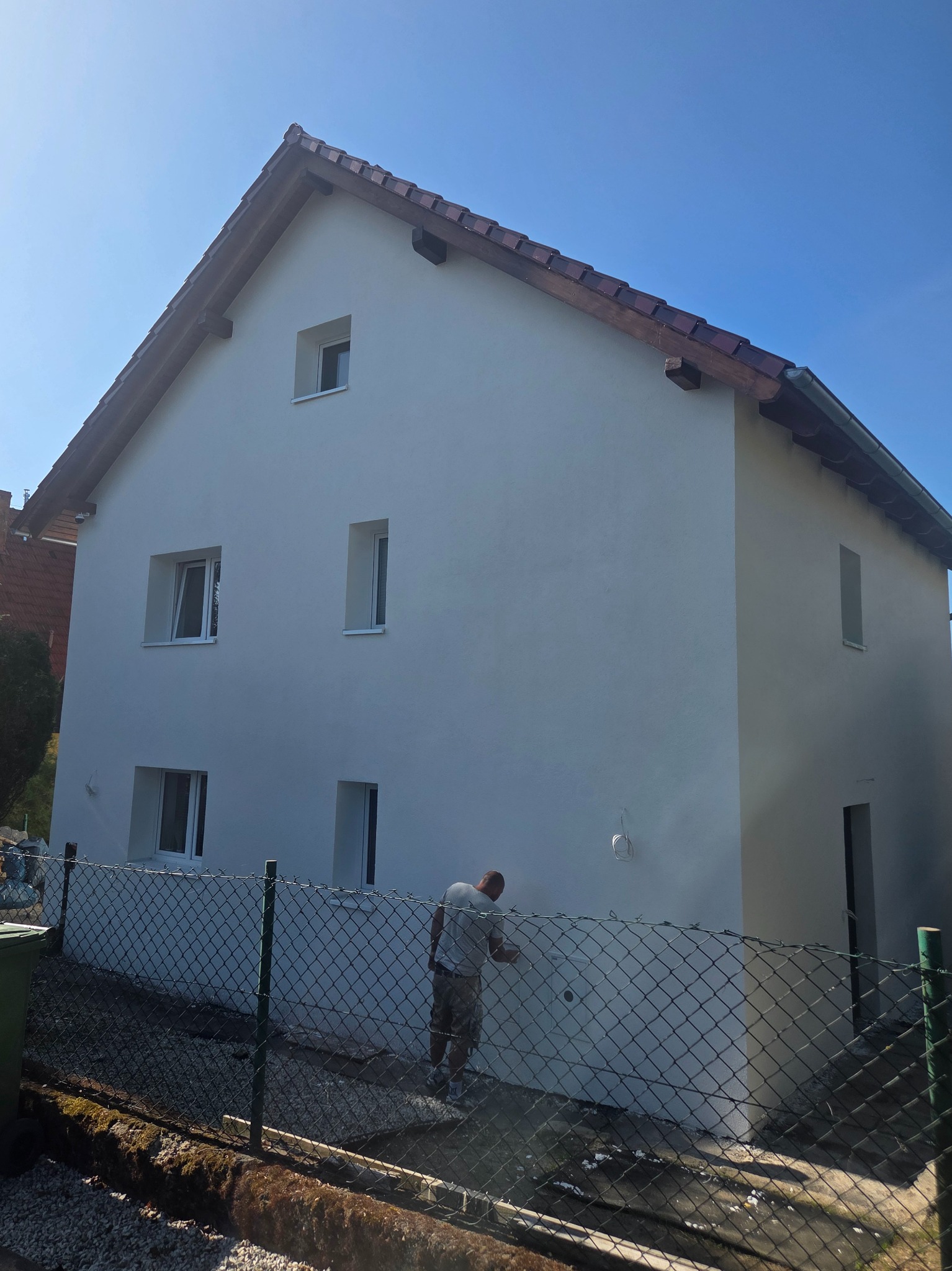 Biały dom jednorodzinny z czerwoną dachówką w trakcie malowania elewacji przez pracownika, widok zza siatki ogrodzeniowej w słoneczny dzień.