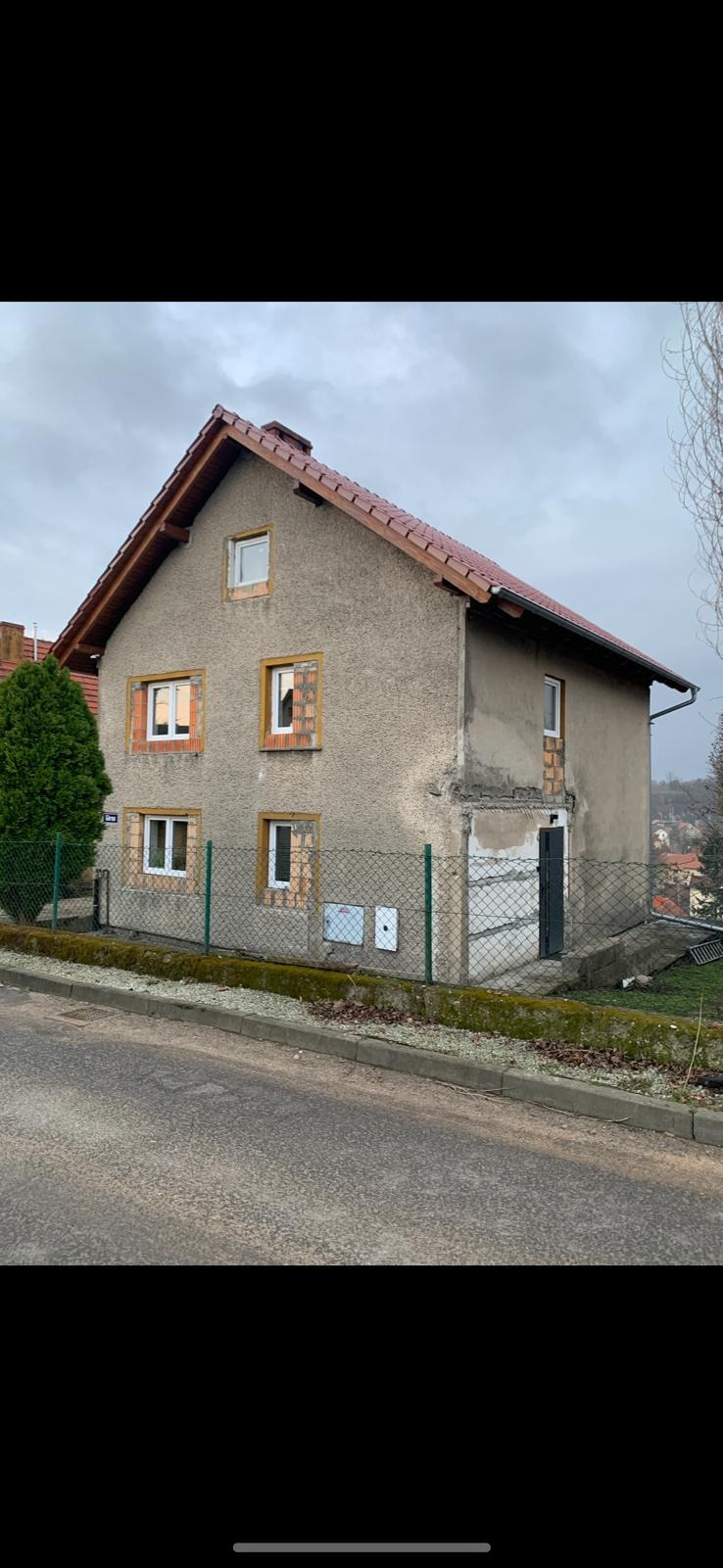 Dwukondygnacyjny dom z częściowo otynkowaną elewacją, nową dachówką i oknami w trakcie montażu, otoczony zielenią i ogrodzeniem z siatki. Widok z poziomu ulicy.