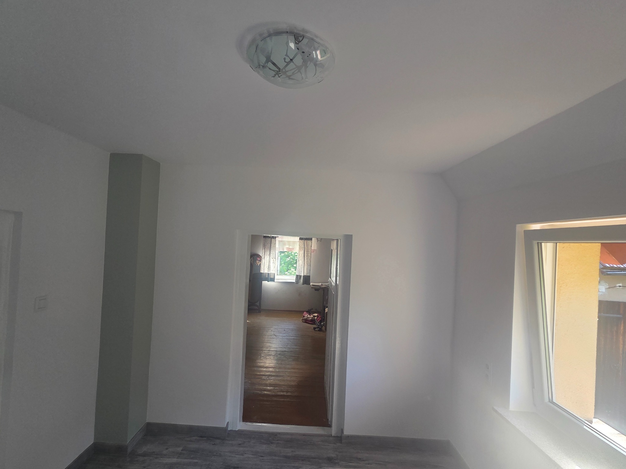 Wnętrze po remoncie: białe ściany, szara kolumna, widok na sąsiedni pokój z drewnianą podłogą, nowoczesna lampa sufitowa, okno z widokiem na zewnątrz.