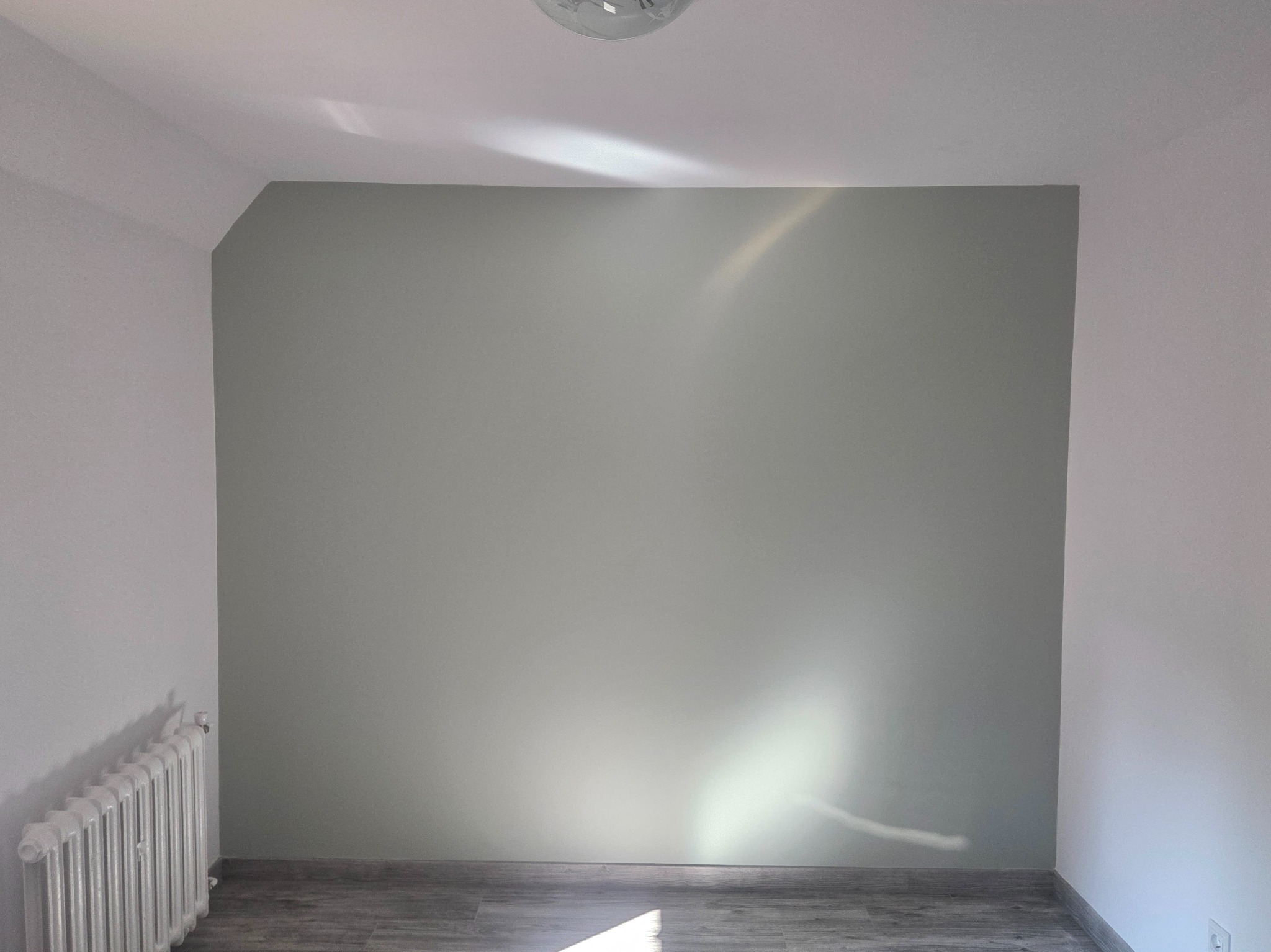 Pomieszczenie po malowaniu: szara ściana z delikatnym połyskiem światła, białe ściany boczne, grzejnik i panele podłogowe. Widok na odnowione wnętrze.