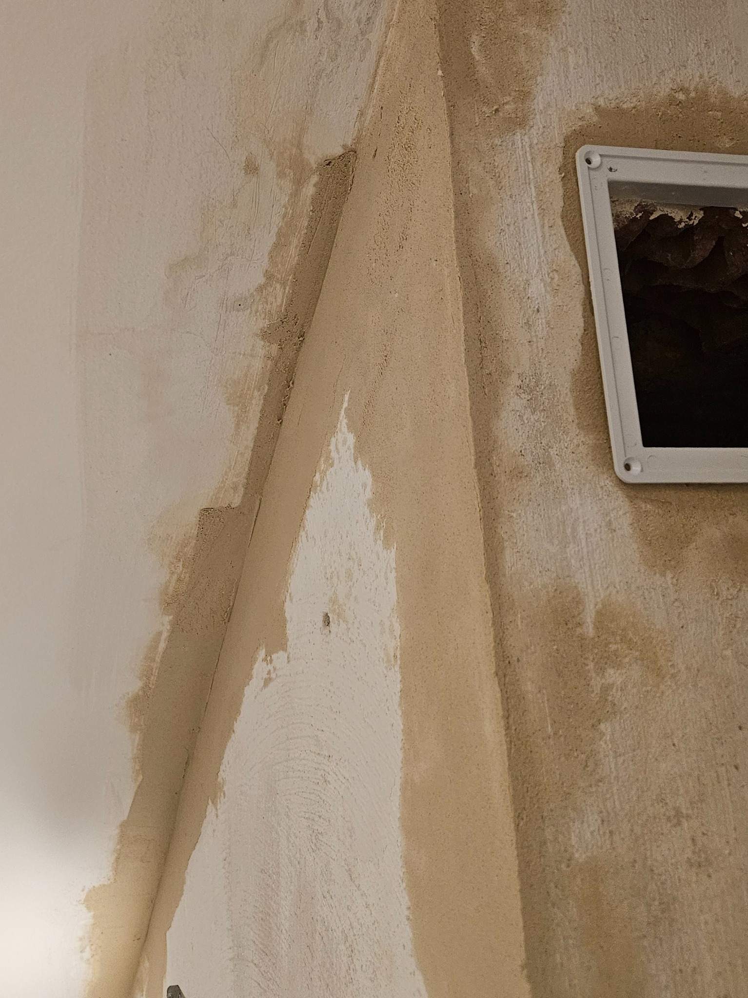 Narożnik pokoju w trakcie prac wykończeniowych, widoczne nałożone warstwy gładzi, fragment kratki wentylacyjnej. Ściany w odcieniach beżu i bieli.