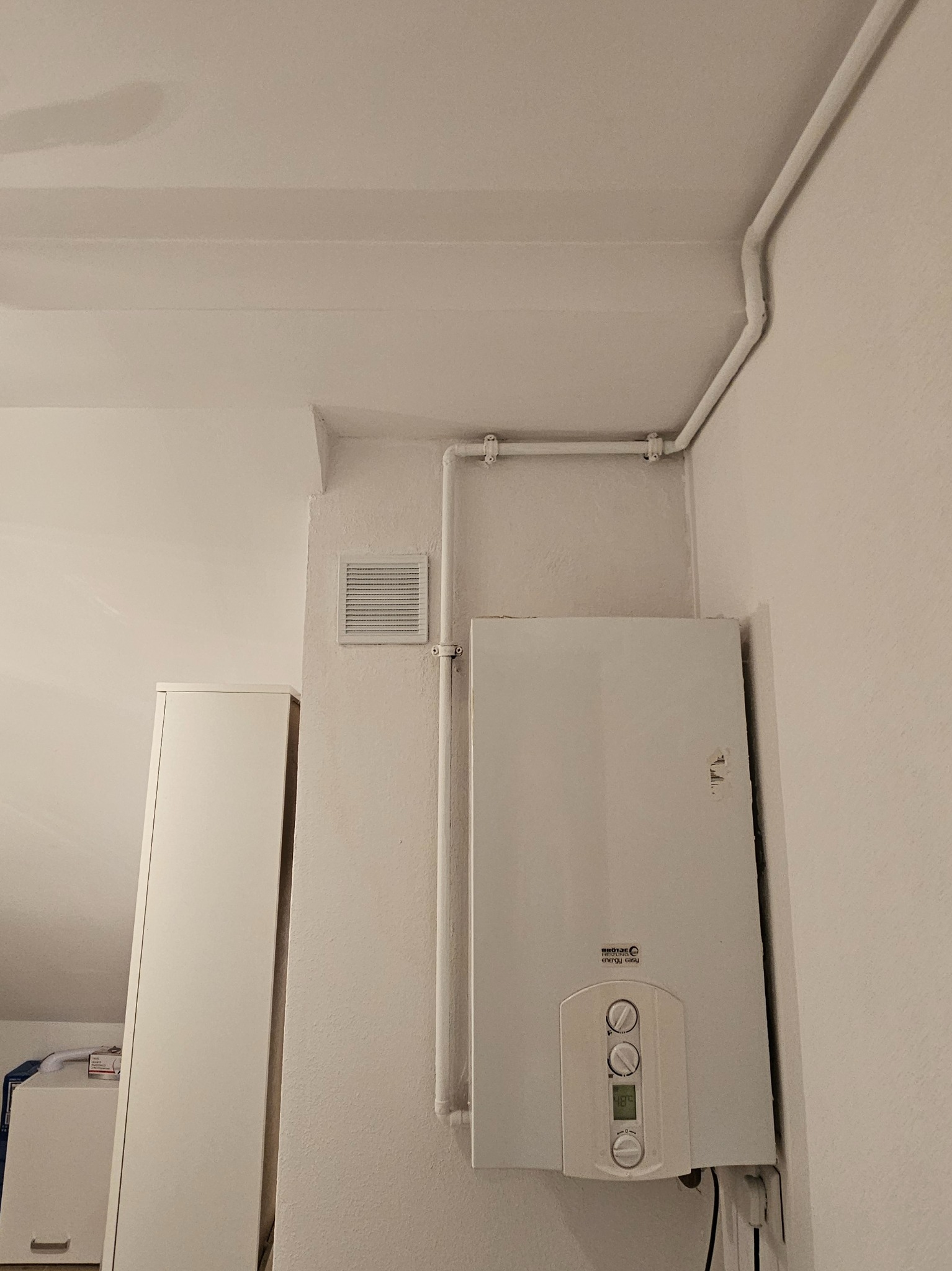 Biały piec gazowy zamontowany na ścianie w rogu pomieszczenia, z widocznymi rurami i kratką wentylacyjną. Obok szafka, a w tle skos dachu.