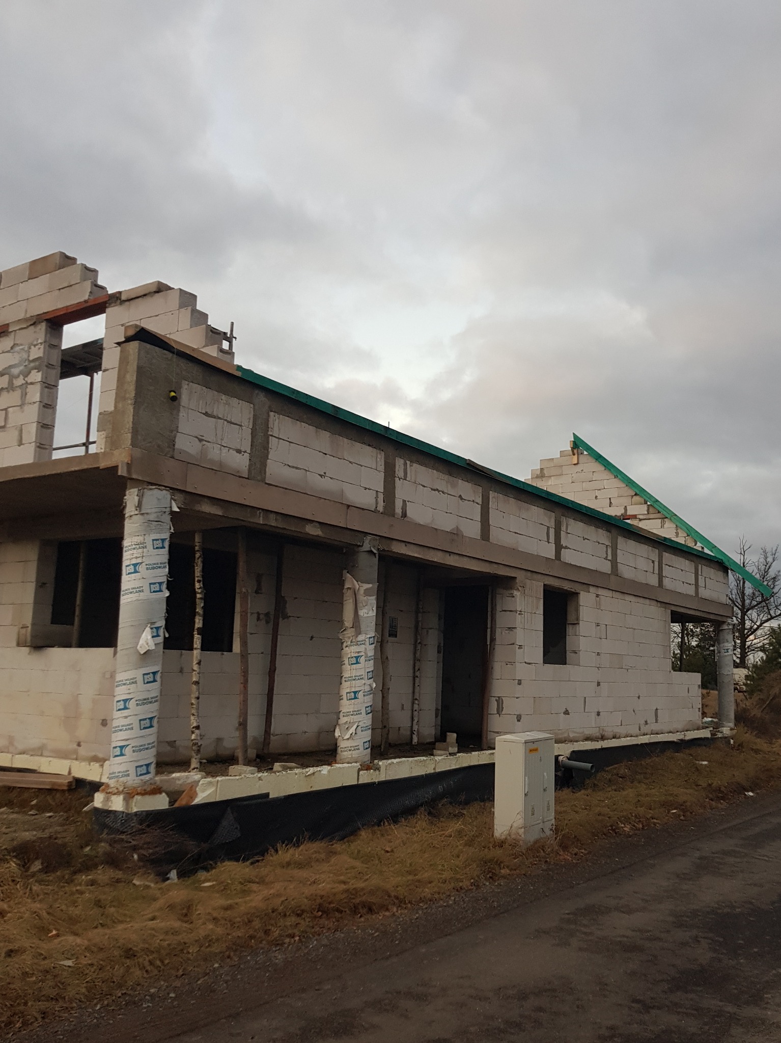 Surowy stan domu w budowie z bloczków, z widocznymi elementami konstrukcyjnymi dachu i izolacją kolumn. Widok na szary budynek w trakcie wznoszenia.