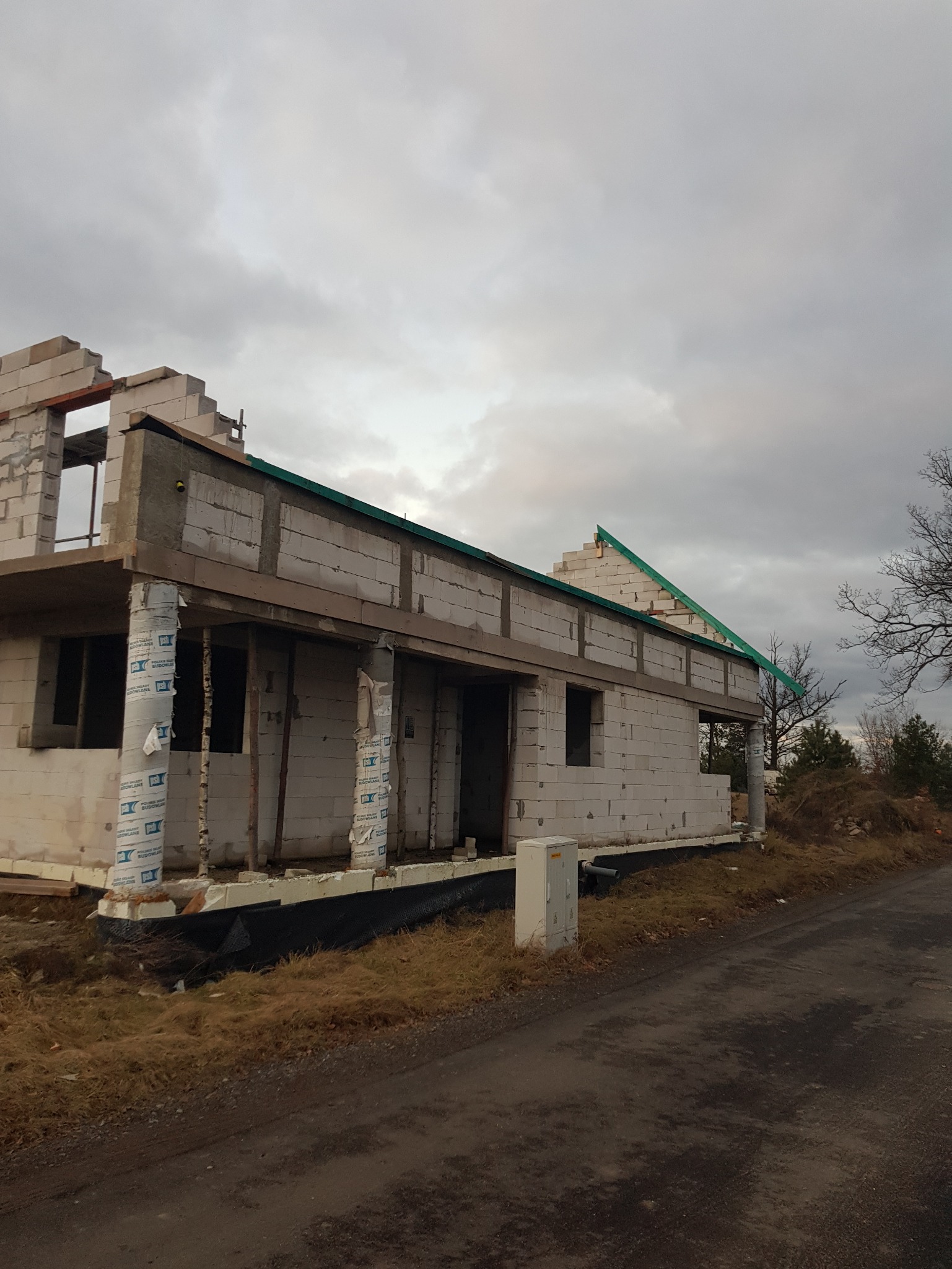 Budowa domu jednorodzinnego w stanie surowym otwartym, widoczne ściany z bloczków, elementy dachu i słupy zabezpieczone folią. Pochmurne niebo w tle.