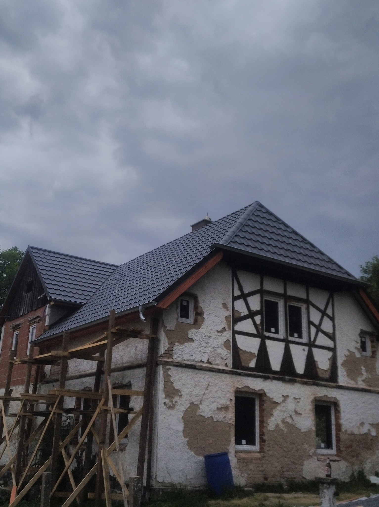 Dom z nowym, ciemnym dachem i widocznymi rusztowaniami. Elewacja w trakcie renowacji, z odsłoniętą cegłą i fragmentami starego tynku. Pochmurne niebo w tle.
